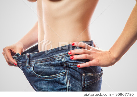 Closeup of slim waist of young woman standing in big jeans showing successful weight loss, indoor studio shot, isolated on light gray background, diet concept. 90907780