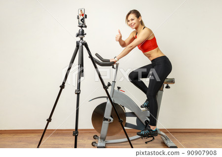 Athletic woman recording tutorial video on cell phone on tripod, riding exercise bike and showing thumb up, wearing sports tights and red top. Indoor studio shot on gray wall background. Athletic woman recording tutorial video on cell phone on tripod, riding exercise bike and showing thumb up, wearing sports tights and red top. Indoor studio shot on gray wall background. 90907908