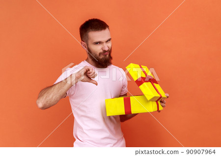 Portrait of bearded man unpacking present to know what he received, being frustrated and disappointed, showing thumb down, wearing pink T-shirt. Indoor studio shot isolated on orange background 90907964
