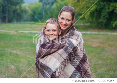 Young sister and brother with freckles on their faces stand covered with plaid in park, get warm, smiling and looking at camera. Young sister and brother with freckles on their faces stand covered with plaid in park, get warm, smiling and looking at camera. 90907967