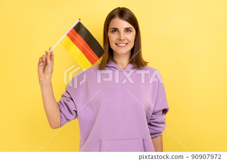 Portrait of satisfied smiling young adult woman standing and waving german flag, celebrating national holiday, wearing purple hoodie. Indoor studio shot isolated on yellow background. 90907972