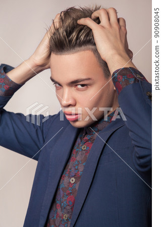 Handsome young fashion model with highlighted undercut haircut posing in blue suit in studio looking with passion at camera and touch and holding his hair. Handsome young fashion model with highlighted undercut haircut posing in blue suit in studio looking with passion at camera and touch and holding his hair. 90908045