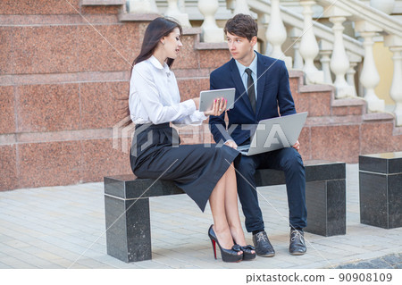 Boss in blue suit and his assistant in white blouse and black skirt discussing their work outdoors. 90908109