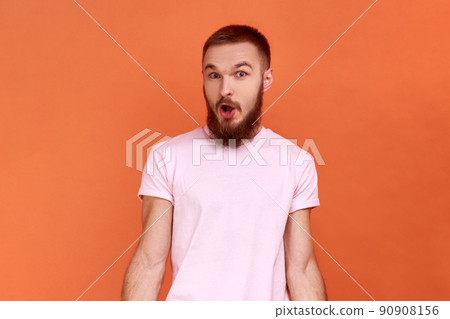 Portrait of amazed bearded man standing with mouth open in surprise, has shocked expression, hears unbelievable news, wearing pink T-shirt. Indoor studio shot isolated on orange background. 90908156