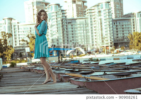 Beautiful young fashion model in green turquois dress and fashion makeup and hairstyle is posing on pier, posing and looking at camera on city background. Beautiful young fashion model in green turquois dress and fashion makeup and hairstyle is posing on pier, posing and looking at camera on city background. 90908261