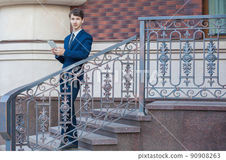 Young handsome businessman standing at steps in the architecture background, holding tablet and looking at camera. Young handsome businessman standing at steps in the architecture background, holding tablet and looking at camera. 90908263