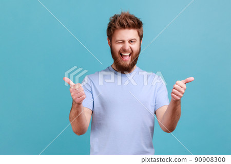 Portrait of bearded man approves incredible promo keeps thumbs up likes and agrees being satisfied with something, winking to camera. Indoor studio shot isolated on blue background. 90908300