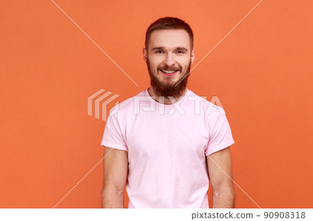 Portrait of happy bearded man looking at camera and smiling, expressing positive emotions, being in good mood, wearing pink T-shirt. Indoor studio shot isolated on orange background. Portrait of happy bearded man looking at camera and smiling, expressing positive emotions, being in good mood, wearing pink T-shirt. Indoor studio shot isolated on orange background. 90908318
