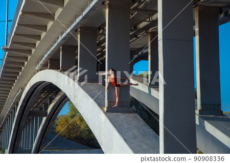 Beautiful young strong healthy woman ballerina gymnast in red leotard posing as model on bridge girder and blue sky and looking into the distance. Beautiful young strong healthy woman ballerina gymnast in red leotard posing as model on bridge girder and blue sky and looking into the distance. 90908336