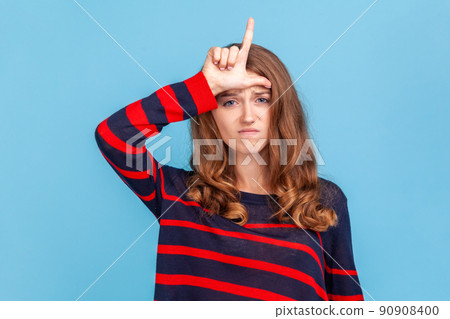 Unhappy depressed woman wearing striped casual style sweater, showing loser gesture, L finger sign on forehead, upset about dismissal, unlucky day.. Indoor studio shot isolated on blue background. Unhappy depressed woman wearing striped casual style sweater, showing loser gesture, L finger sign on forehead, upset about dismissal, unlucky day.. Indoor studio shot isolated on blue background. 90908400