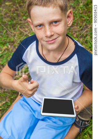 Cute young caucasian kid with freckles on his face in blue shorts and white and blue T-shirt and tablet and working in park and smiling. top view, looking at camera thumbs up. Cute young caucasian kid with freckles on his face in blue shorts and white and blue T-shirt and tablet and working in park and smiling. top view, looking at camera thumbs up. 90908415