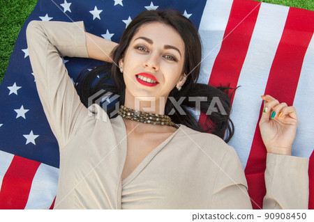 Beautiful sexy young woman with classic dress lying down on american flag in the park. fashion model holding us smiling and looking at camera. USA lifestyle with toothy smile. 90908450