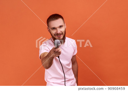 Portrait of bearded man posing with microphone in hands, offers mic, journalist asking questions, looking at camera, wearing pink T-shirt. Indoor studio shot isolated on orange background. Portrait of bearded man posing with microphone in hands, offers mic, journalist asking questions, looking at camera, wearing pink T-shirt. Indoor studio shot isolated on orange background. 90908459