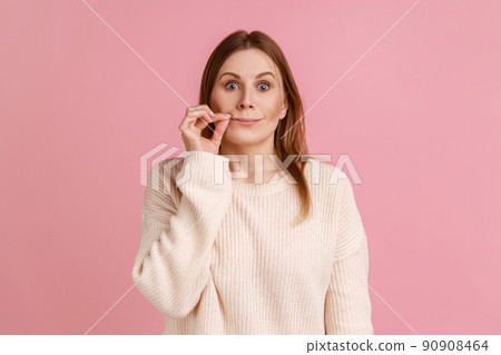 I won't tell anyone. Intimidated blond woman zipping lips and looking at camera, covering mouth promising to keep terrible secret, wearing white sweater. Indoor studio shot isolated on pink background I won't tell anyone. Intimidated blond woman zipping lips and looking at camera, covering mouth promising to keep terrible secret, wearing white sweater. Indoor studio shot isolated on pink background 90908464