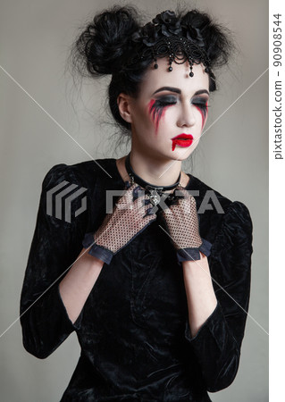 Young beautiful gothic woman with white skin and red lips with bloody drops wearing black collar with spikes. Red smokey eyes. Halloween makeup. Young beautiful gothic woman with white skin and red lips with bloody drops wearing black collar with spikes. Red smokey eyes. Halloween makeup. 90908544