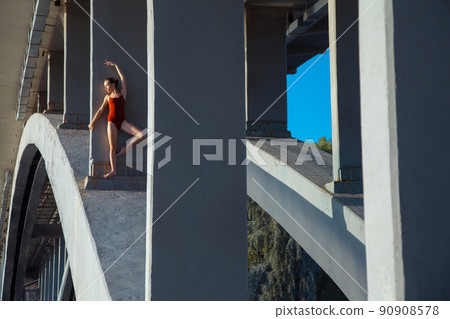 Beautiful young strong healthy woman ballerina gymnast in red leotard posing as model on bridge girder and blue sky and looking into the distance. 90908578