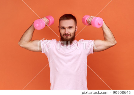 Portrait of strong athletic handsome young bearded man holding rose dumbbells in raised arms, healthy lifestyle, wearing pink T-shirt. Indoor studio shot isolated on orange background. Portrait of strong athletic handsome young bearded man holding rose dumbbells in raised arms, healthy lifestyle, wearing pink T-shirt. Indoor studio shot isolated on orange background. 90908645