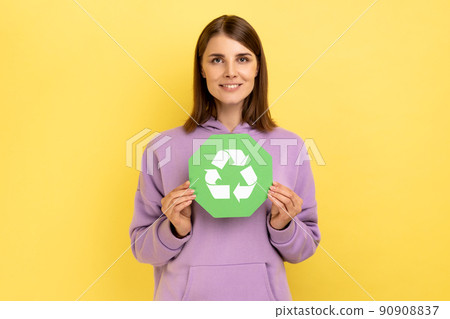 Smiling woman with dark hair holding in hands green recycling sing, ecology concept, looking at camera, wearing purple hoodie. Indoor studio shot isolated on yellow background. 90908837