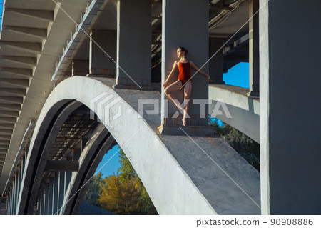 Beautiful young strong healthy woman ballerina gymnast in red leotard posing as model on bridge girder and blue sky and looking into the distance. 90908886