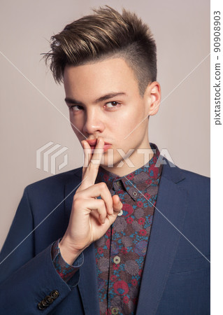 Handsome young fashion model with highlighted undercut haircut posing in blue suit in studio looking with passion at camera with finger on his lips. Handsome young fashion model with highlighted undercut haircut posing in blue suit in studio looking with passion at camera with finger on his lips. 90908903
