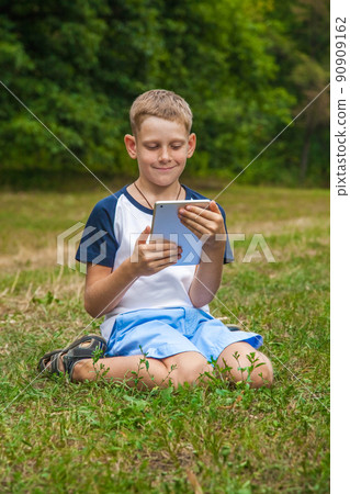 Cute young caucasian kid with freckles on his face in blue shorts and white and blue T-shirt and tablet and working in park and smiling. sitting and looking at display with smile. Cute young caucasian kid with freckles on his face in blue shorts and white and blue T-shirt and tablet and working in park and smiling. sitting and looking at display with smile. 90909162