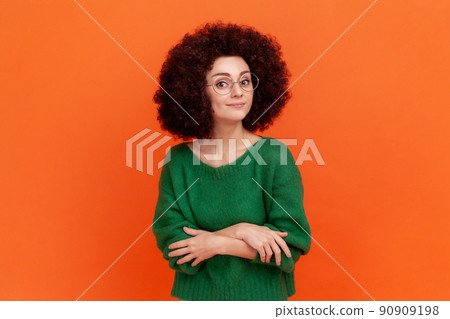 Portrait of confident woman with Afro hairstyle wearing green casual style sweater and optical glasses, standing with folded arms, looking at camera. Indoor studio shot isolated on orange background. Portrait of confident woman with Afro hairstyle wearing green casual style sweater and optical glasses, standing with folded arms, looking at camera. Indoor studio shot isolated on orange background. 90909198