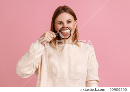 Portrait of positive attractive blond woman standing, holding magnifying glass on his teeth, looking at camera, wearing white sweater. Indoor studio shot isolated on pink background. 90909200