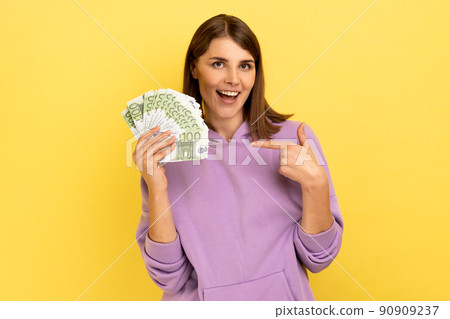 Amazed smiling woman pointing at euro banknotes in her hand, having optimistic positive expression, big profit, wearing purple hoodie. Indoor studio shot isolated on yellow background. Amazed smiling woman pointing at euro banknotes in her hand, having optimistic positive expression, big profit, wearing purple hoodie. Indoor studio shot isolated on yellow background. 90909237