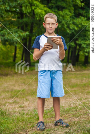 Cute young caucasian kid with freckles on his face in blue shorts and white and blue T-shirt and tablet and working in park and smiling. standing and looking at camera. 90909328