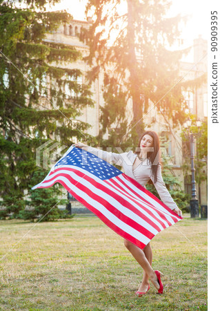 American flag and woman (4th july). Beautiful young woman with classic dress holding american flag in the park. fashion model holding us smiling and looking at camera. usa lifestyle.. 90909395