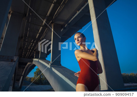 Beautiful young strong healthy woman ballerina gymnast in red leotard posing as model on bridge girder and blue sky and looking into the distance. 90909457