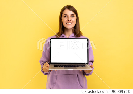 Positive optimistic woman standing showing laptop with white empty screen for promotion, looking at camera with smile, wearing purple hoodie. Indoor studio shot isolated on yellow background. Positive optimistic woman standing showing laptop with white empty screen for promotion, looking at camera with smile, wearing purple hoodie. Indoor studio shot isolated on yellow background. 90909508