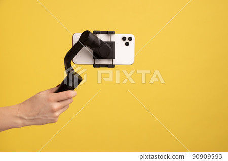 Closeup side view of woman hand holding steadicam with phone, for making video or has livestream. Indoor studio shot isolated on yellow background. Closeup side view of woman hand holding steadicam with phone, for making video or has livestream. Indoor studio shot isolated on yellow background. 90909593