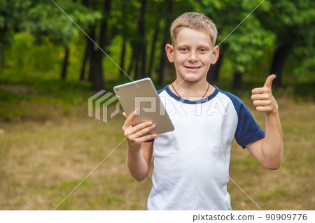 Cute young caucasian kid with freckles on his face in blue shorts and white and blue T-shirt and tablet and working in park and smiling. looking at camera, smiling with thumb up. Cute young caucasian kid with freckles on his face in blue shorts and white and blue T-shirt and tablet and working in park and smiling. looking at camera, smiling with thumb up. 90909776