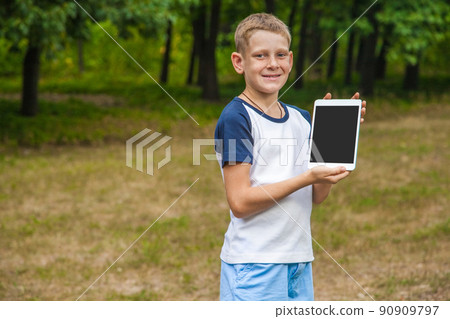 Cute young caucasian kid with freckles on his face in blue shorts and white and blue T-shirt and tablet and working in park and smiling. looking at camera with smile and showing display. Cute young caucasian kid with freckles on his face in blue shorts and white and blue T-shirt and tablet and working in park and smiling. looking at camera with smile and showing display. 90909797