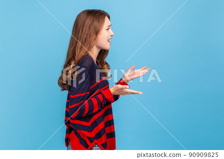 Side view of unsatisfied annoyed woman wearing striped casual style sweater standing with raised hands in anger and shouting why how, what do you want. Indoor studio shot isolated on blue background. 90909825