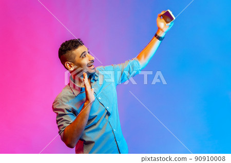 Portrait of friendly man in shirt talking on video call and waving hello gesture, having online conversation on mobile phone. Indoor studio shot isolated on colorful neon light background. 90910008