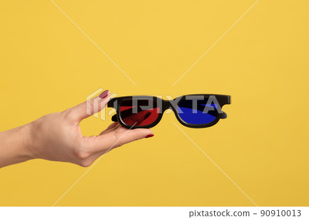 Profile side view closeup of woman hand holding and showing 3d eyeglasses for watching movie in cinema. Indoor studio shot isolated on yellow background. Profile side view closeup of woman hand holding and showing 3d eyeglasses for watching movie in cinema. Indoor studio shot isolated on yellow background. 90910013