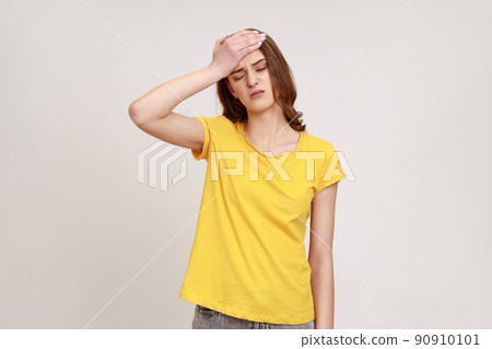 Portrait of upset beautiful female of young age in yellow T- shirt standing with facepalm gesture, blaming himself, feeling regret because of bad memory. Indoor studio shot isolated on gray background 90910101