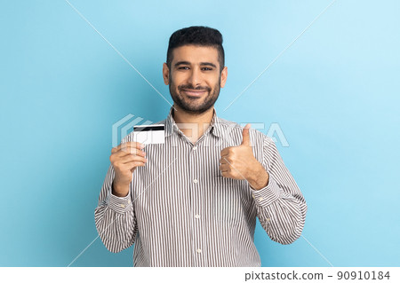 Businessman holding credit card, looking with toothy smile, recommend bank or shopping discounts, showing thumb up, wearing striped shirt. Indoor studio shot isolated on blue background. 90910184