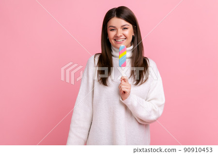 Smiling satisfied brunette female holding delicious ice cream, looks at camera with happy expression, wearing white casual style sweater. Indoor studio shot isolated on pink background. 90910453