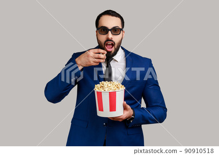Impressed bearded man getting experience VR headset, using augmented reality eyeglasses being in virtual reality, watching movie with popcorn. Indoor studio shot isolated on gray background. 90910518