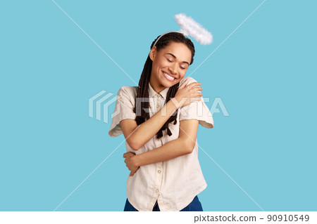 I love myself. Angelic woman with dreadlocks and saint nimbus embracing herself and smiling with pleasure, feeling complacency, wearing white shirt. Indoor studio shot isolated on blue background. 90910549