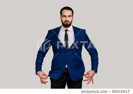 Portrait of sad unhappy bearded man showing empty pockets inside out, going bankrupt, no cash for living, wearing official style suit. Indoor studio shot isolated on gray background. Portrait of sad unhappy bearded man showing empty pockets inside out, going bankrupt, no cash for living, wearing official style suit. Indoor studio shot isolated on gray background. 90910617