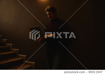 Confident portrait of a serious handsome mature Caucasian businessman, entrepreneur, copywriter working online on laptop standing against a dark wall lit with dim light near stairs in office interior Confident portrait of a serious handsome mature Caucasian businessman, entrepreneur, copywriter working online on laptop standing against a dark wall lit with dim light near stairs in office interior 90911947