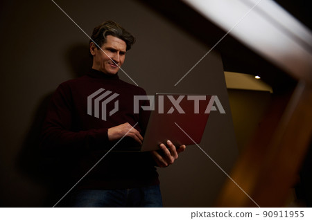 Attractive handsome mature European man working remotely on laptop against dark brown wall background. Businessman, entrepreneur, copywriter, journalist freelance. Online work concept Attractive handsome mature European man working remotely on laptop against dark brown wall background. Businessman, entrepreneur, copywriter, journalist freelance. Online work concept 90911955
