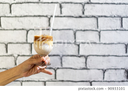 Coffee Macchiato and milk layer with ice in champagne glass was held by Asian grandmother hand in front of white brick wall wallpaper. Coffee Macchiato and milk layer with ice in champagne glass was held by Asian grandmother hand in front of white brick wall wallpaper. 90933101