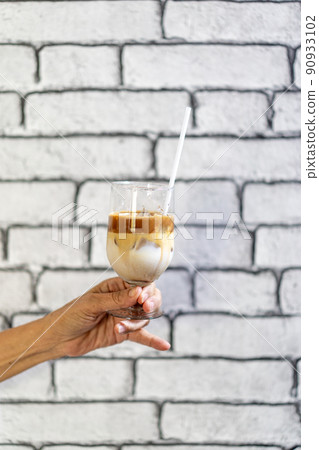 Coffee Macchiato and milk layer with ice in champagne glass was held by Asian grandmother hand in front of white brick wall wallpaper. 90933102