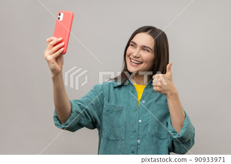 Satisfied attractive woman showing thumb up while communicating by video call or streaming, taking selfie on smartphone, wearing casual jacket. Indoor studio shot isolated on gray background. 90933971