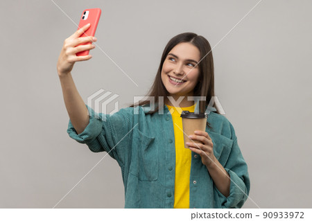 Portrait of attractive young adult woman posing and making selfie photo with smile, blogger lifestyle, wearing casual style jacket. Indoor studio shot isolated on gray background. 90933972